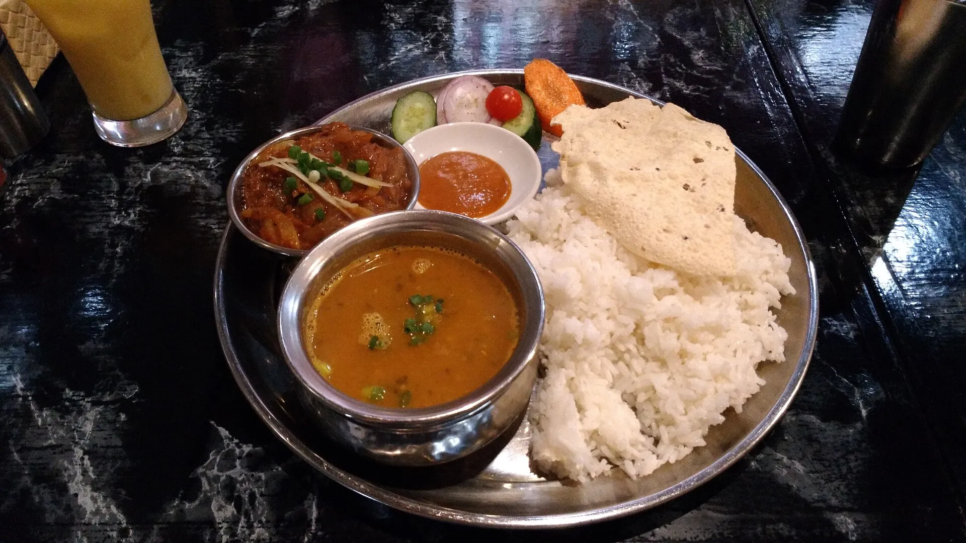 A dal bhat set on a steel plate with rice, lentil soup, curry, pickle, papad and a glass of juice